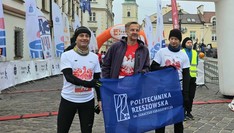 Trzech biegaczy z flagą z napisem Politechnika Rzeszowska na tle banera reklamowego